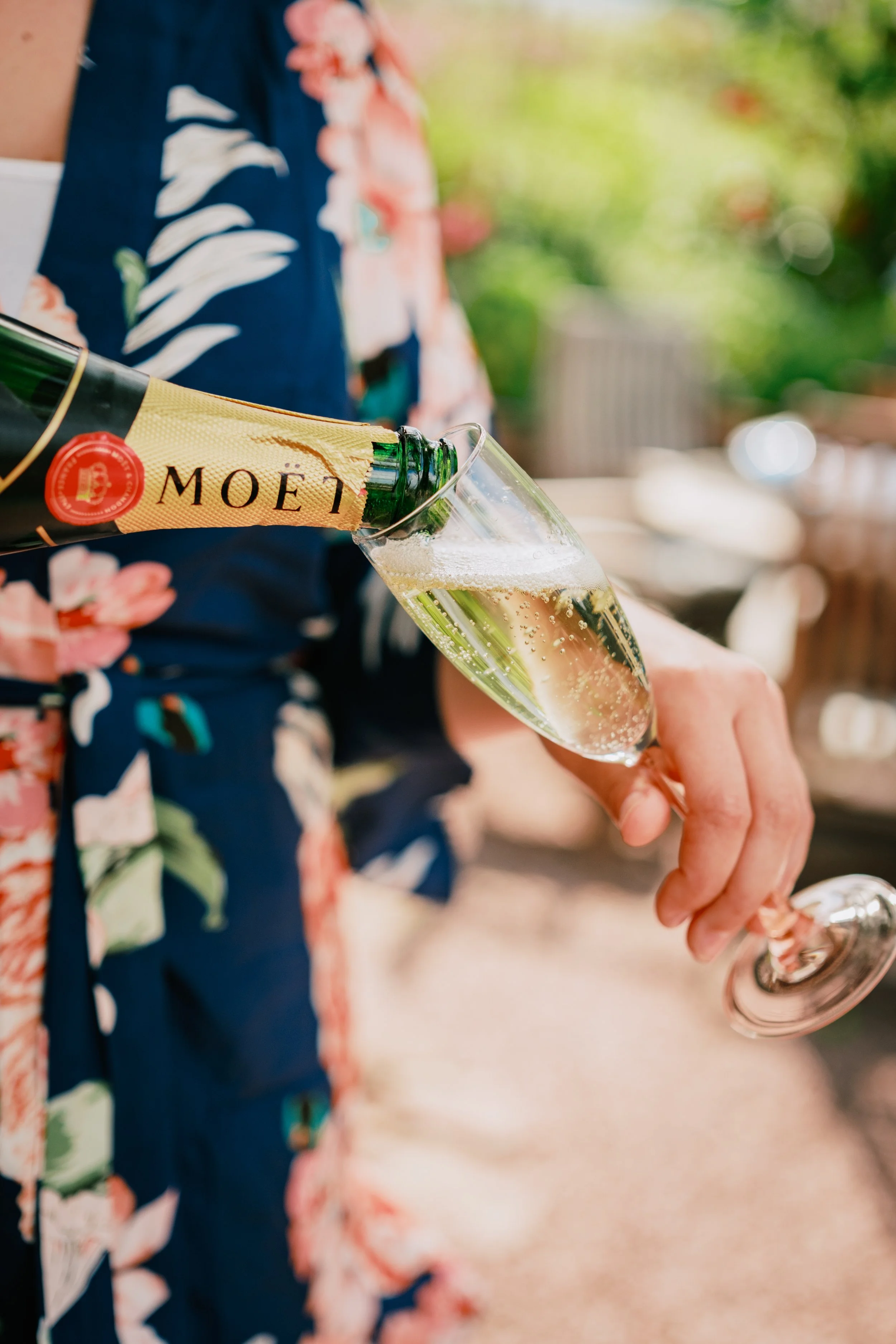 Person in a floral dress pours champagne from a bottle into a flute glass during a celebration or gathering.