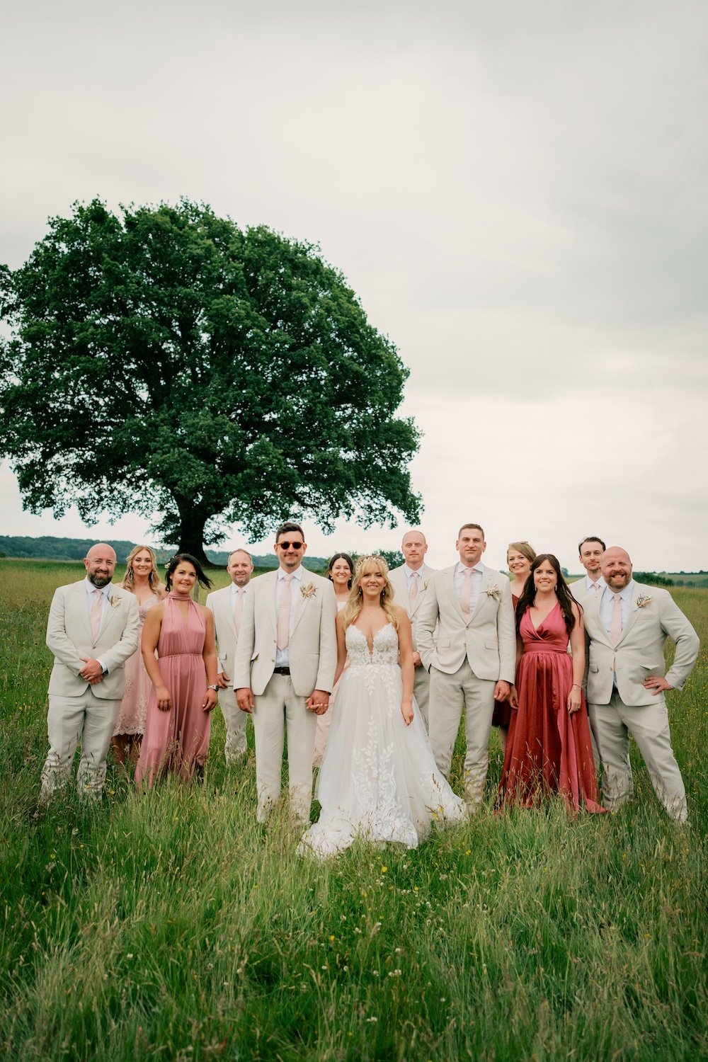 Group of people dressed in wedding attire standing in a grassy field with a large tree in the background.