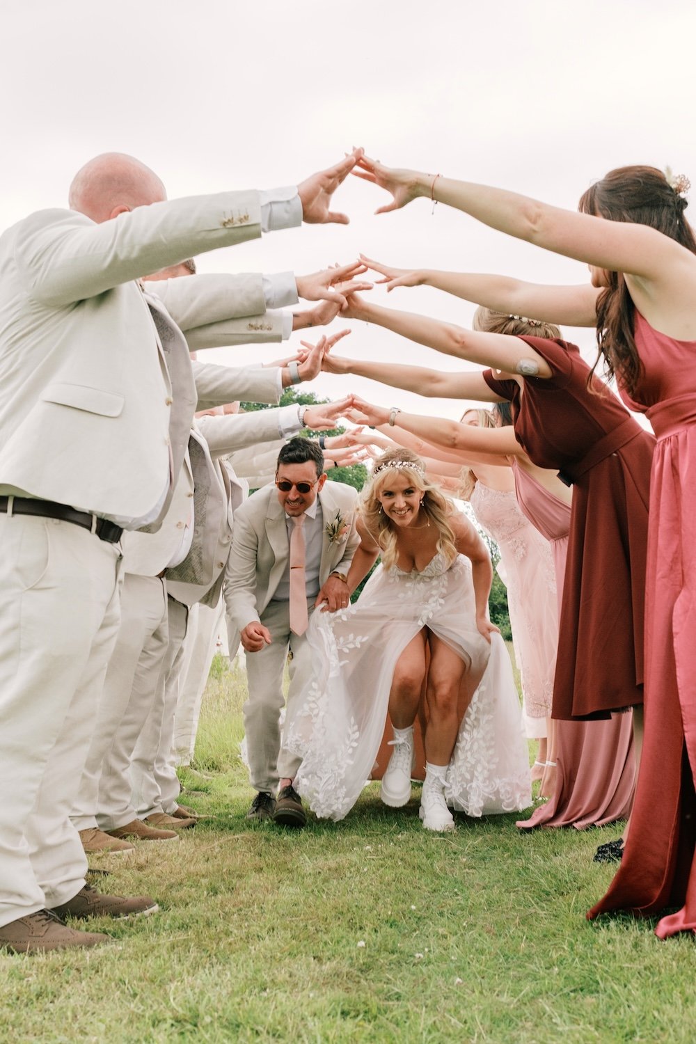 A newlywed couple walking through a tunnel formed by wedding guests who are reaching towards them with outstretched arms, on a grassy outdoor setting.