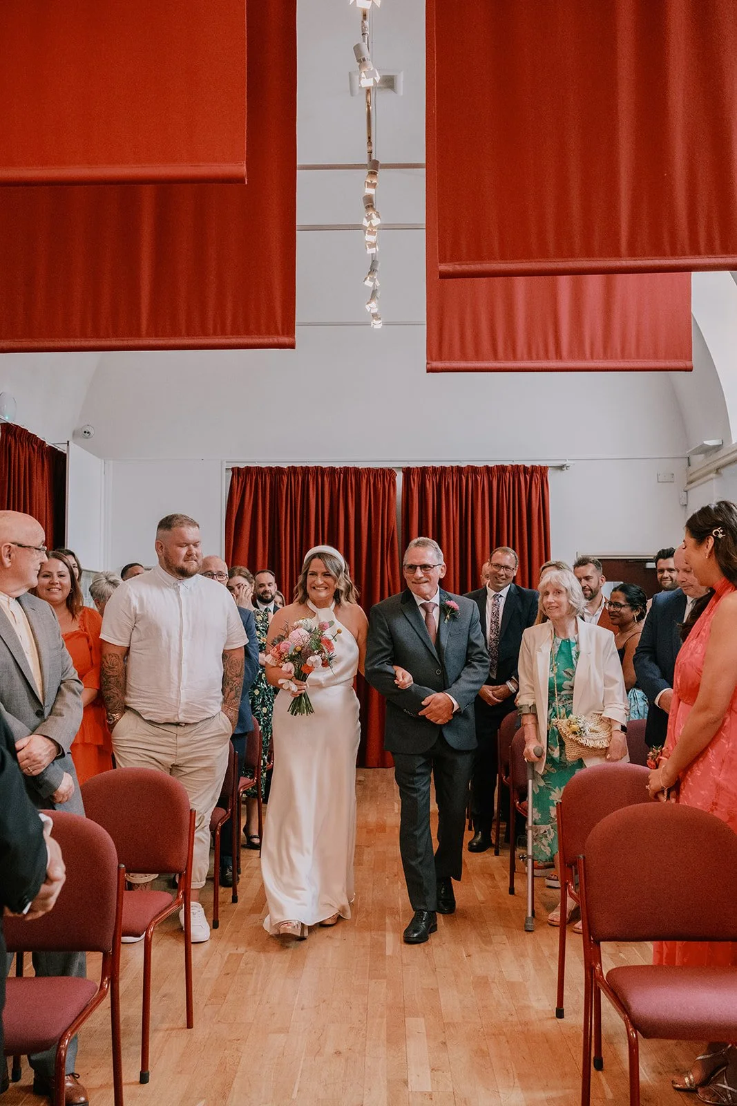 Bride walking down aisle with a man, surrounded by guests in a hall with red curtains.