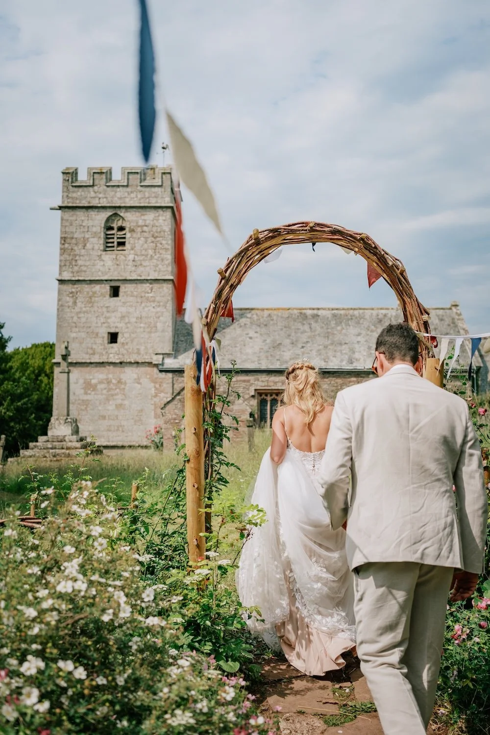 Bride and groom walking through a garden archway towards a stone church with a tower, under a partly cloudy sky.