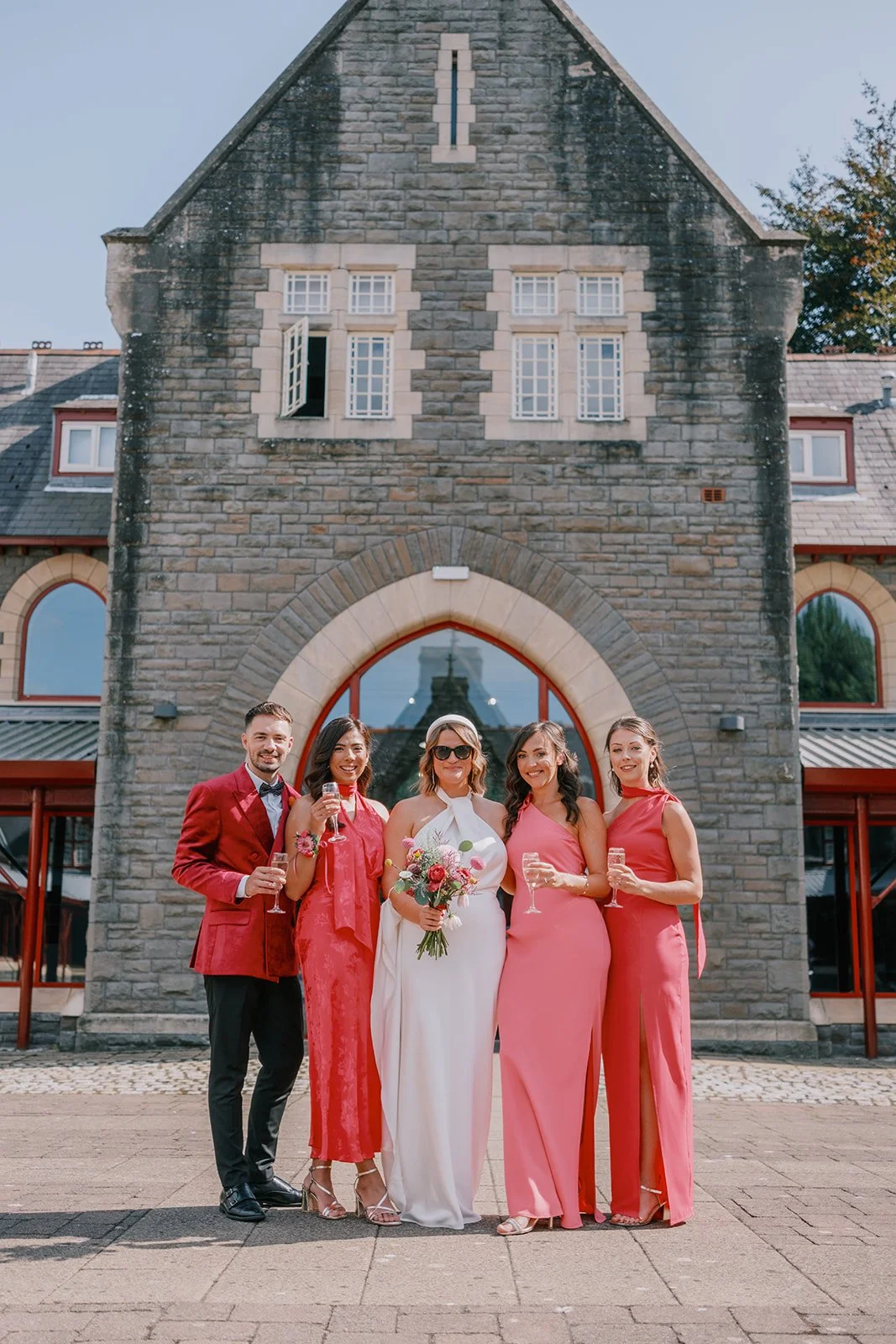Bride with wedding party in front of stone building, all holding champagne glasses, wearing formal attire.