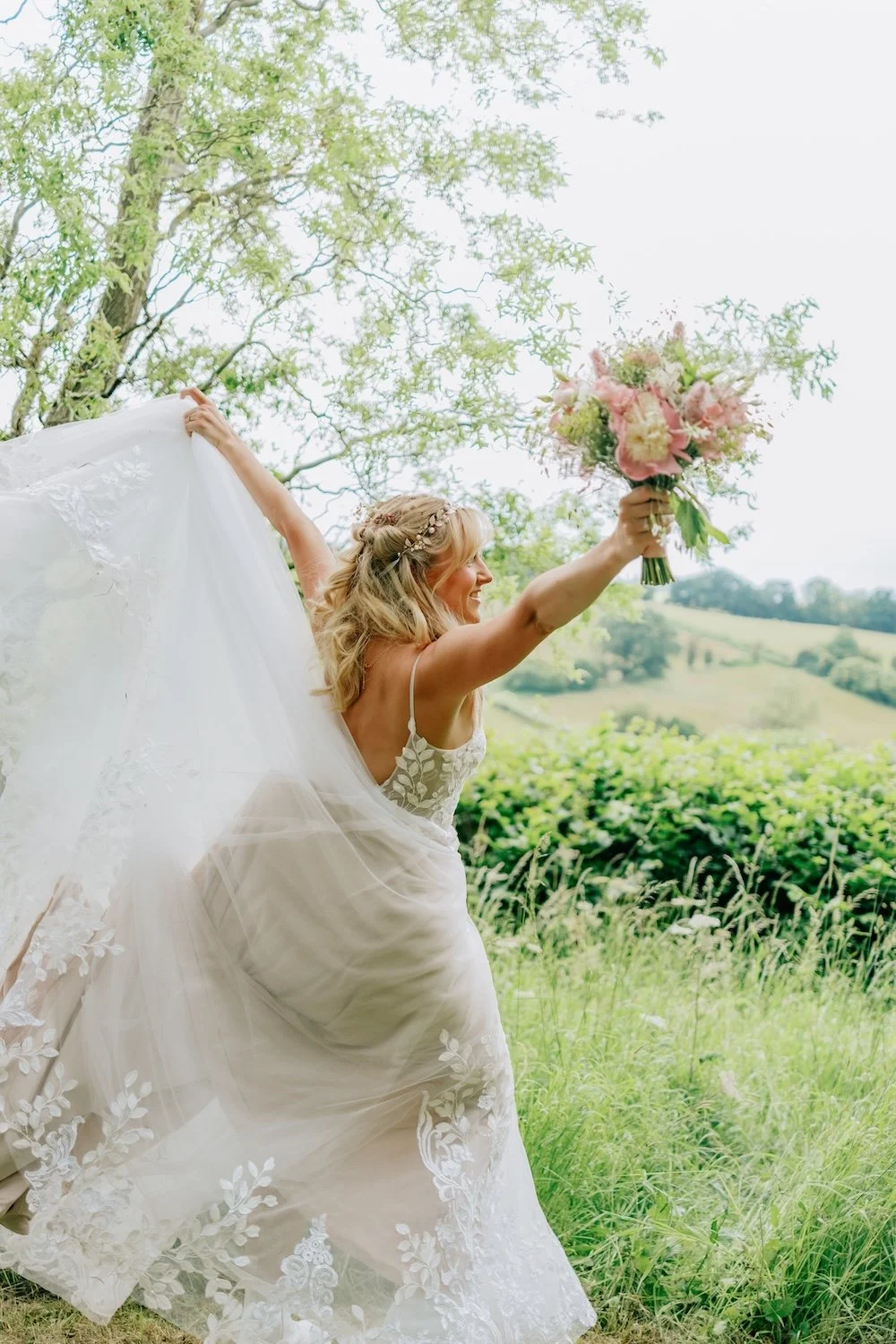 A smiling bride in a white wedding dress holding a bouquet of pink and white flowers in a scenic outdoor setting with greenery and rolling hills.