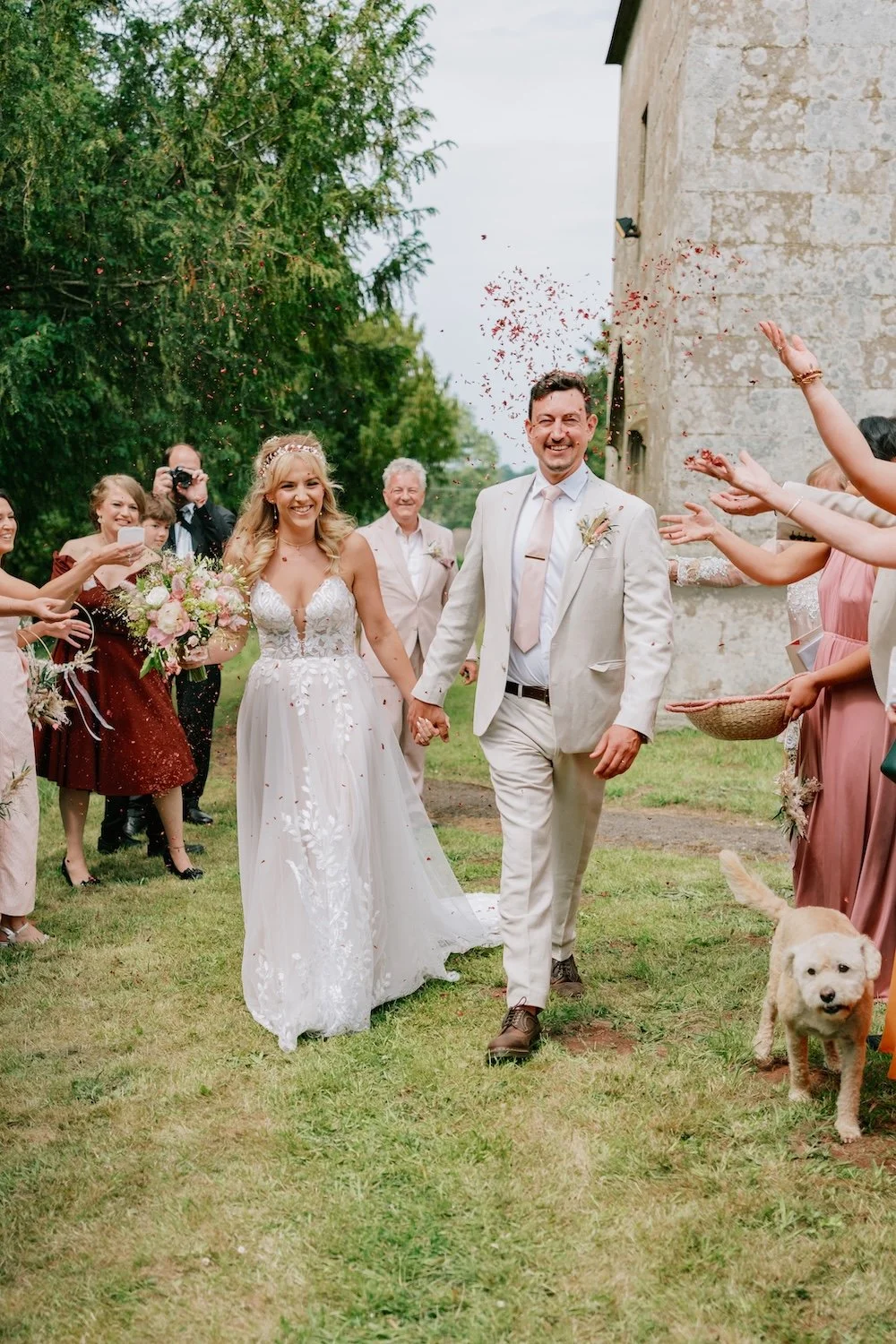 A newlywed couple holding hands and walking outdoors amid smiling friends and family, with one woman holding a bouquet, and confetti in the air near an old stone building and green trees.