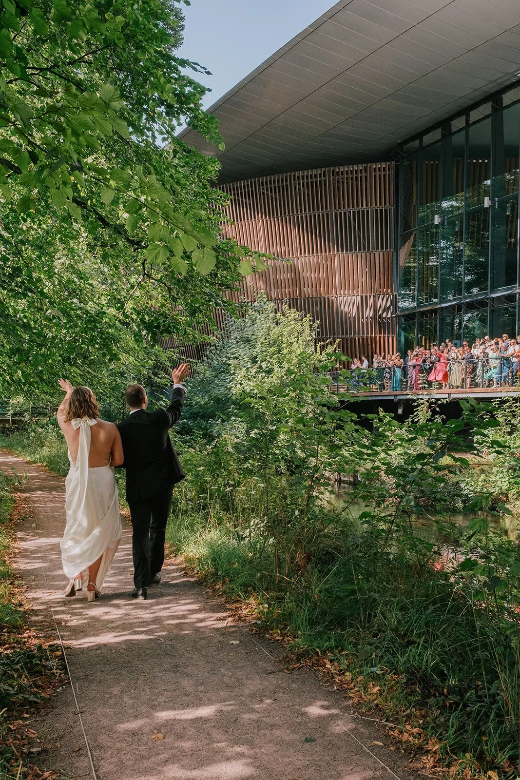 A couple in wedding attire walking along a wooded path, with guests gathered on a balcony of a modern building in the background.