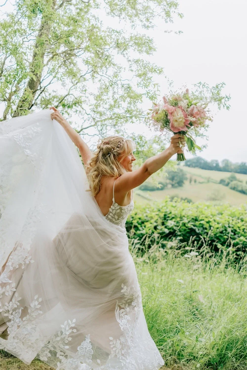 A bride in a white wedding dress holding a bouquet of pink and cream flowers, smiling outdoors in a scenic green landscape with trees and rolling hills.