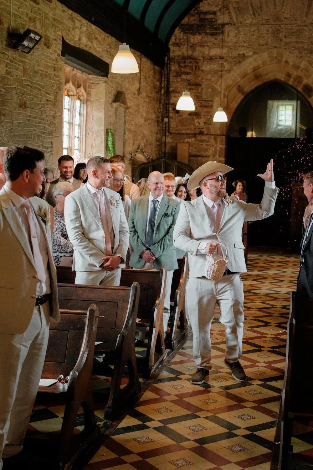 Groom and guests at a wedding ceremony inside a rustic stone church, with one guest dressed in a light suit, cowboy hat and sunglasses, waving to the groom.
