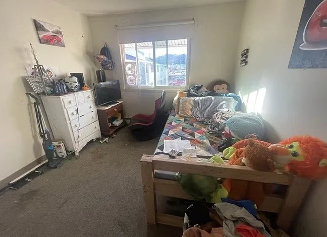 A small bedroom with a twin bed covered in a colorful quilt, stuffed animals, and pillows, a white dresser with a TV and other items, a red and black office chair, and a window showing neighboring buildings and mountains in the distance.