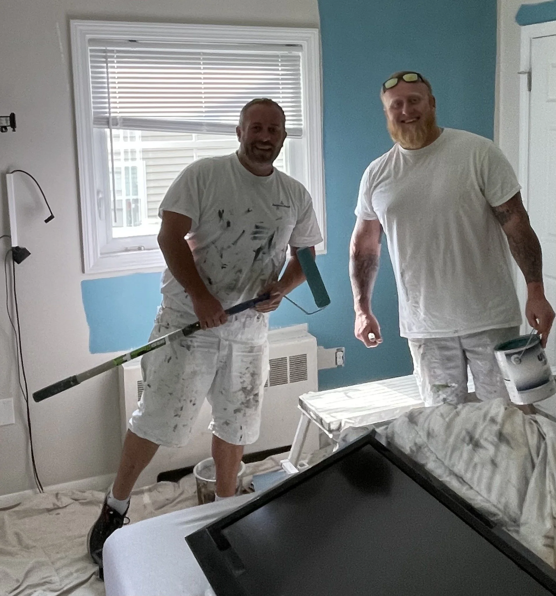 Two men painting a room's wall together, smiling and holding painting tools, with paint and supplies nearby.