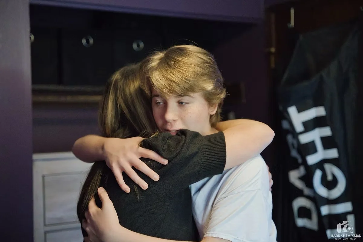 A boy hugging a girl in a room with dark purple walls and a black flag with white text.