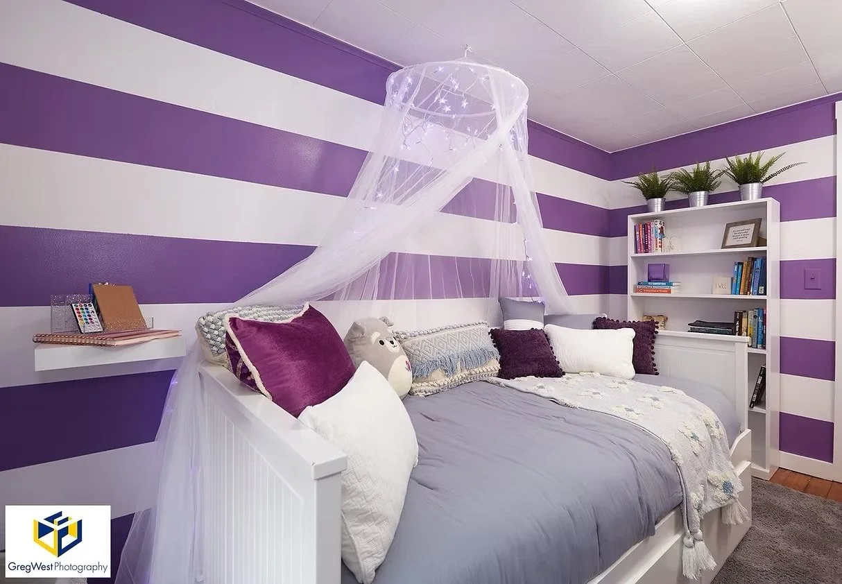 A bedroom with purple and white striped walls, a white bed with multiple pillows, a sheer canopy hanging above, a white bookcase with potted plants and books, and a small wall-mounted shelf with a color wheel and notebooks.