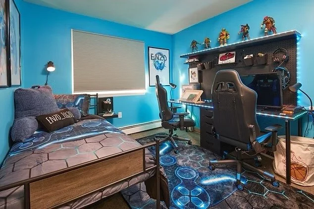 Bedroom with blue walls, bed with hexagon-patterned blanket, desk with gaming setup, and backlit RGB gaming chair, decorated with action figures on a shelf.