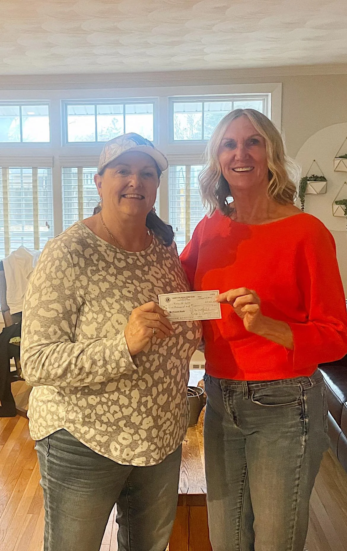 Two women smiling and holding a check together in a bright room with large windows and wooden floors.