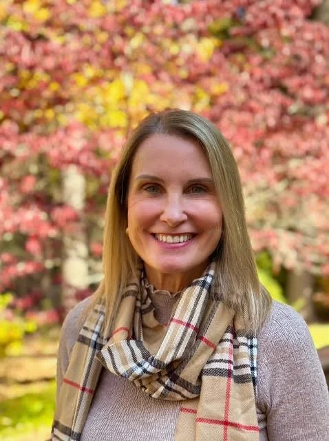 A woman smiling outdoors with pink and yellow autumn leaves in the background