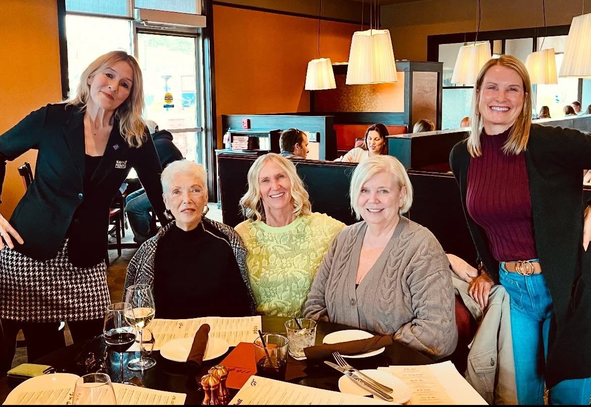 Five women at a restaurant table, smiling and posing for a photo. Two women are standing, three women are seated, and the table has wine glasses, plates, and menus.