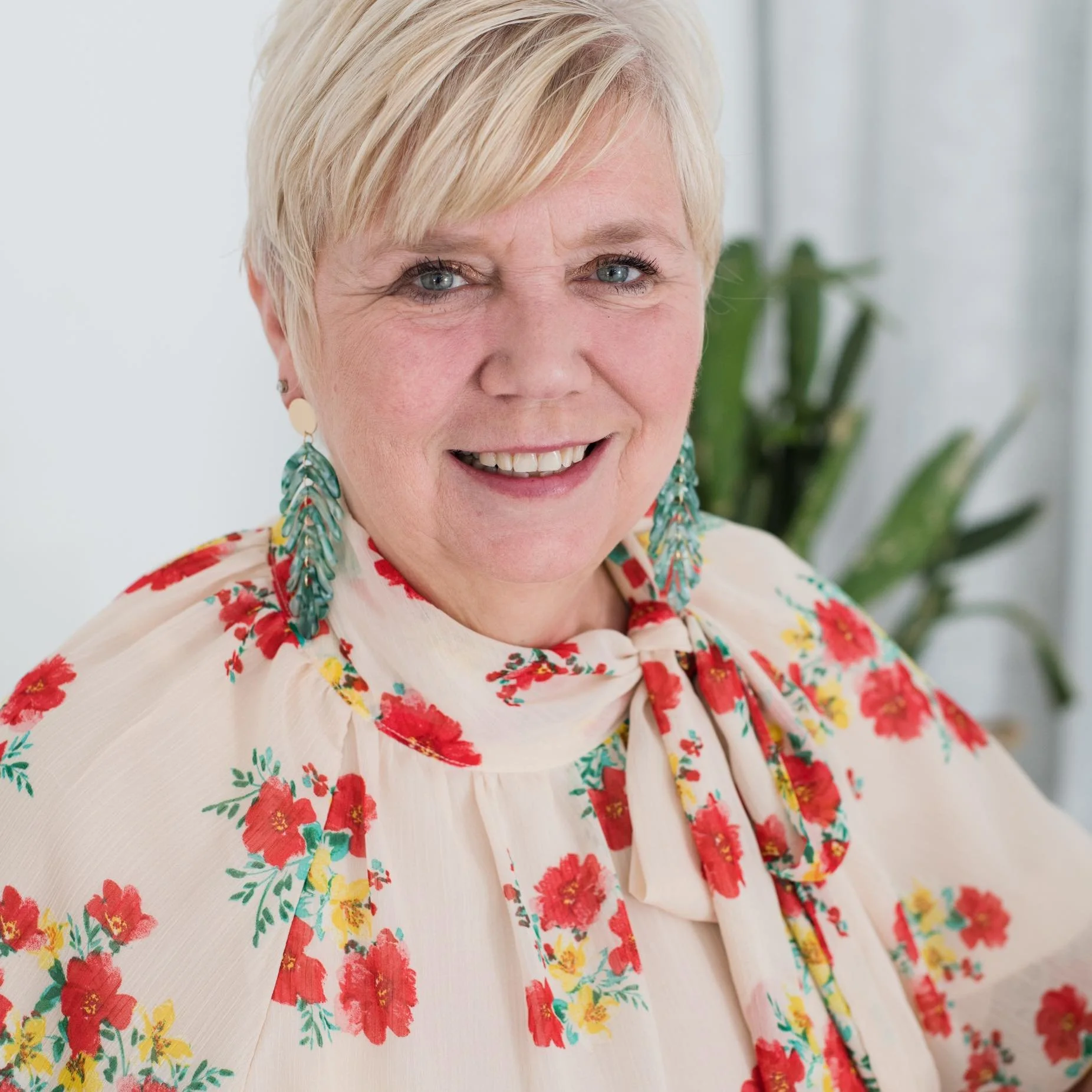 Portrait of an elderly woman with short blonde hair, wearing a floral blouse with red, yellow, and green print, and large teal earrings. She is smiling and has a plant in the background.