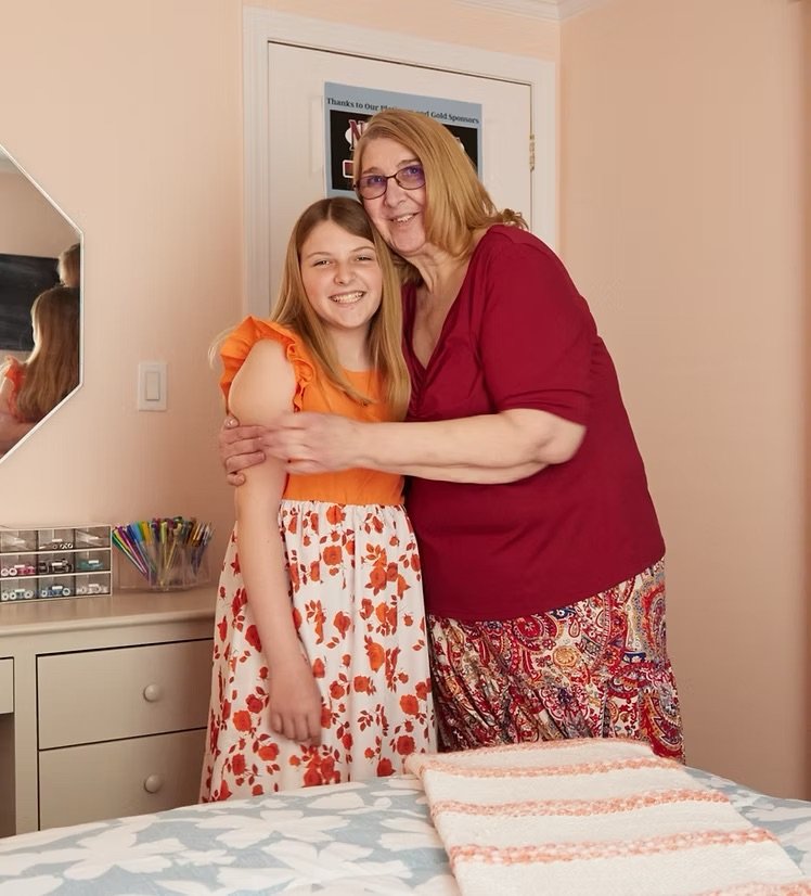 A girl and an older woman hugging and smiling in a bedroom, with a dresser, a mirror, and a bed visible in the background.
