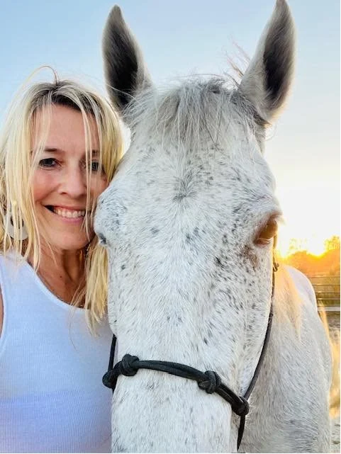 A woman with blonde hair smiling next to a large white horse with spotted markings, outdoor setting at sunset.