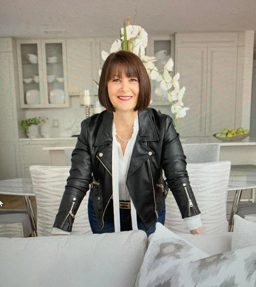 Woman with short brown hair wearing a black leather jacket, white shirt, and necklace, smiling and leaning forward on a couch in a bright dining room with white cabinets, a large white flower arrangement, and green apples on a bowl.