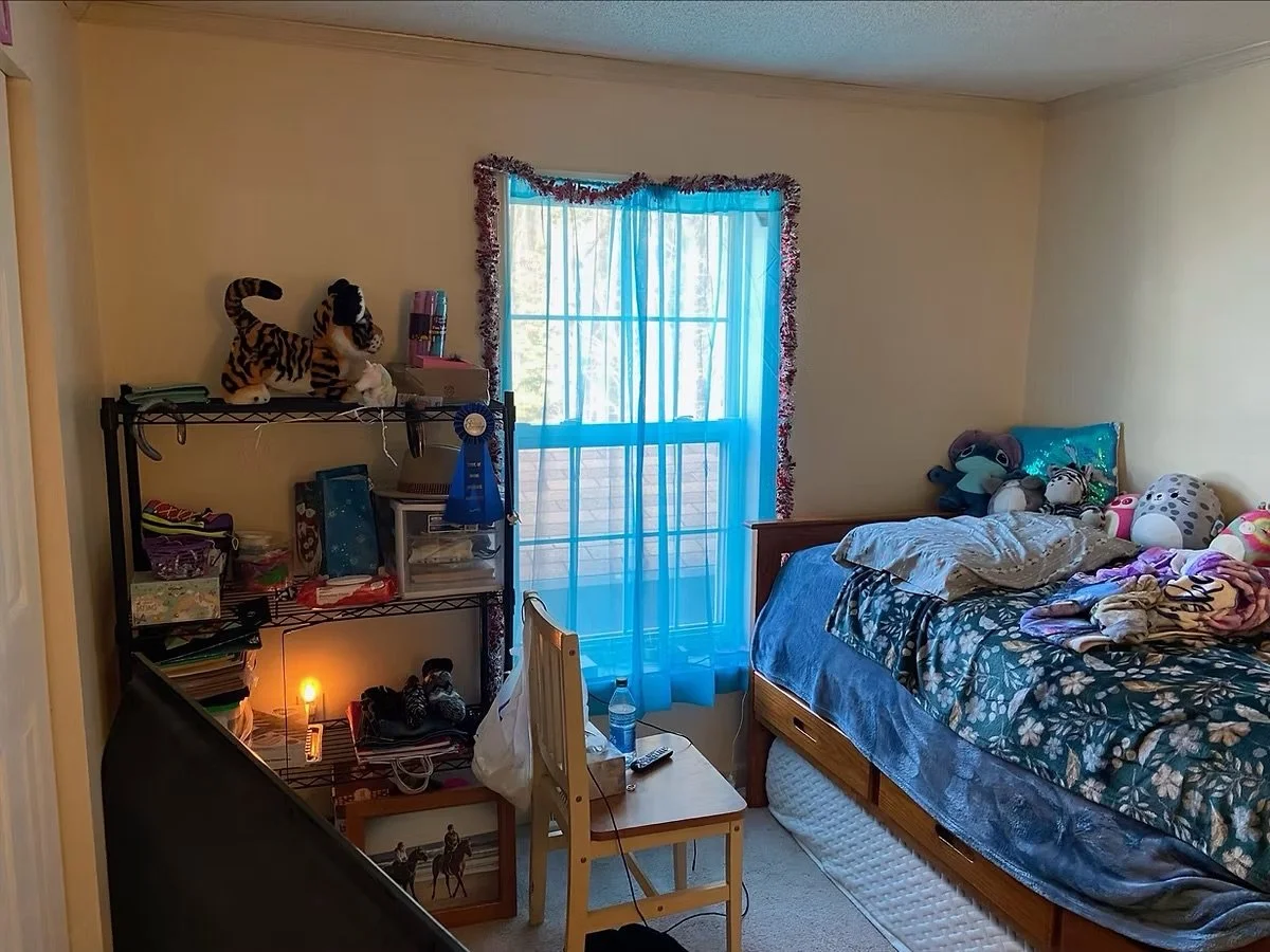 A bedroom with a window dressed in blue curtains, a bed with stuffed animals and pillows, a metal shelf with various items including plush tigers, books, and a blue ribbon, and a small cardboard table with a water bottle and remote.