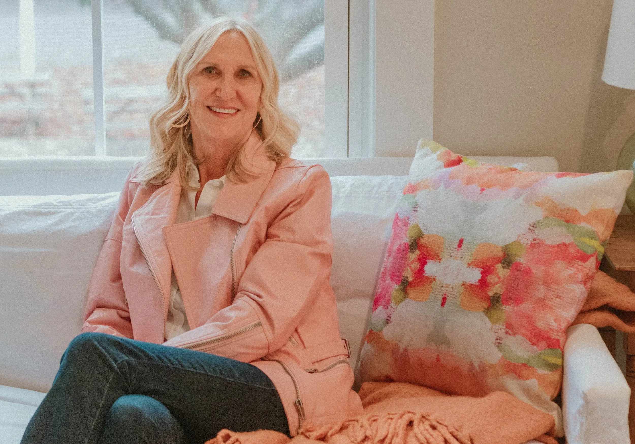 A woman with blonde hair smiling and sitting on a white couch with a colorful pillow, in a room with a large window in the background.