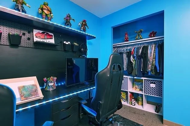 A blue-colored bedroom or gaming room with a black desk, gaming chair, and a closet with hanging clothes. The desk has a computer monitor, colored pencils, and a framed artwork. Wall-mounted shelves above hold action figures and a toy car.