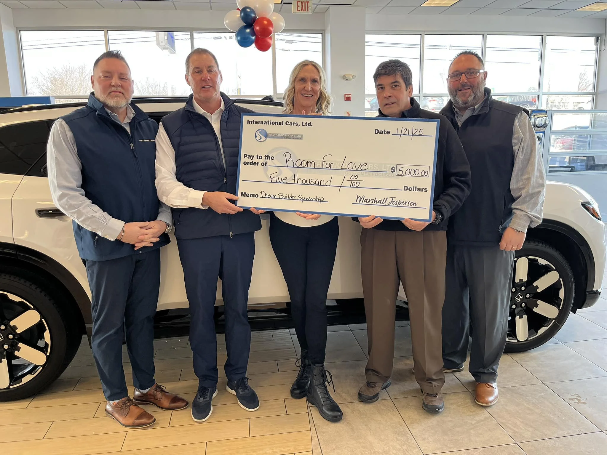 Group of five people standing indoors in front of a car, holding a large check for $5,000 made out to Boom for Love, with balloons in the background.