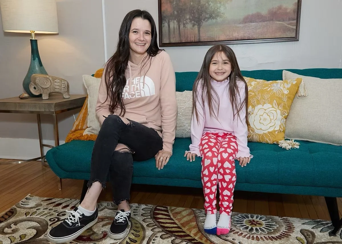 A woman with long dark hair and a girl with long light brown hair sitting on a teal sofa in a living room, smiling at the camera. The woman is wearing a beige hoodie, ripped black jeans, and black sneakers. The girl is wearing pink pajamas with heart patterns and white socks with pink toes. A framed painting of trees is on the wall behind them, and there is a side table with a lamp, a decorative elephant figurine, and a colorful cushion.