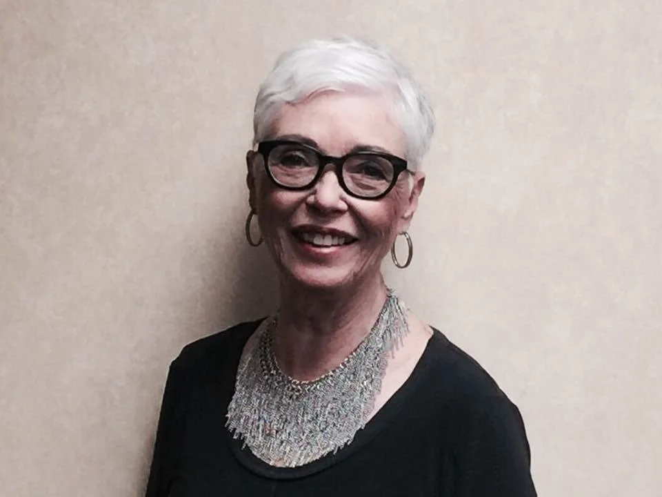 A smiling older woman with short white hair, black glasses, hoop earrings, wearing a silver necklace and a black top against a plain beige wall.