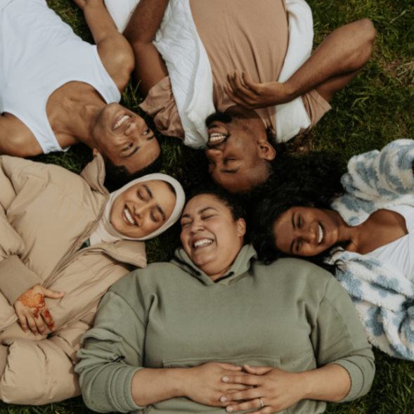 Five diverse friends lying on grass in a circle, smiling and laughing together.