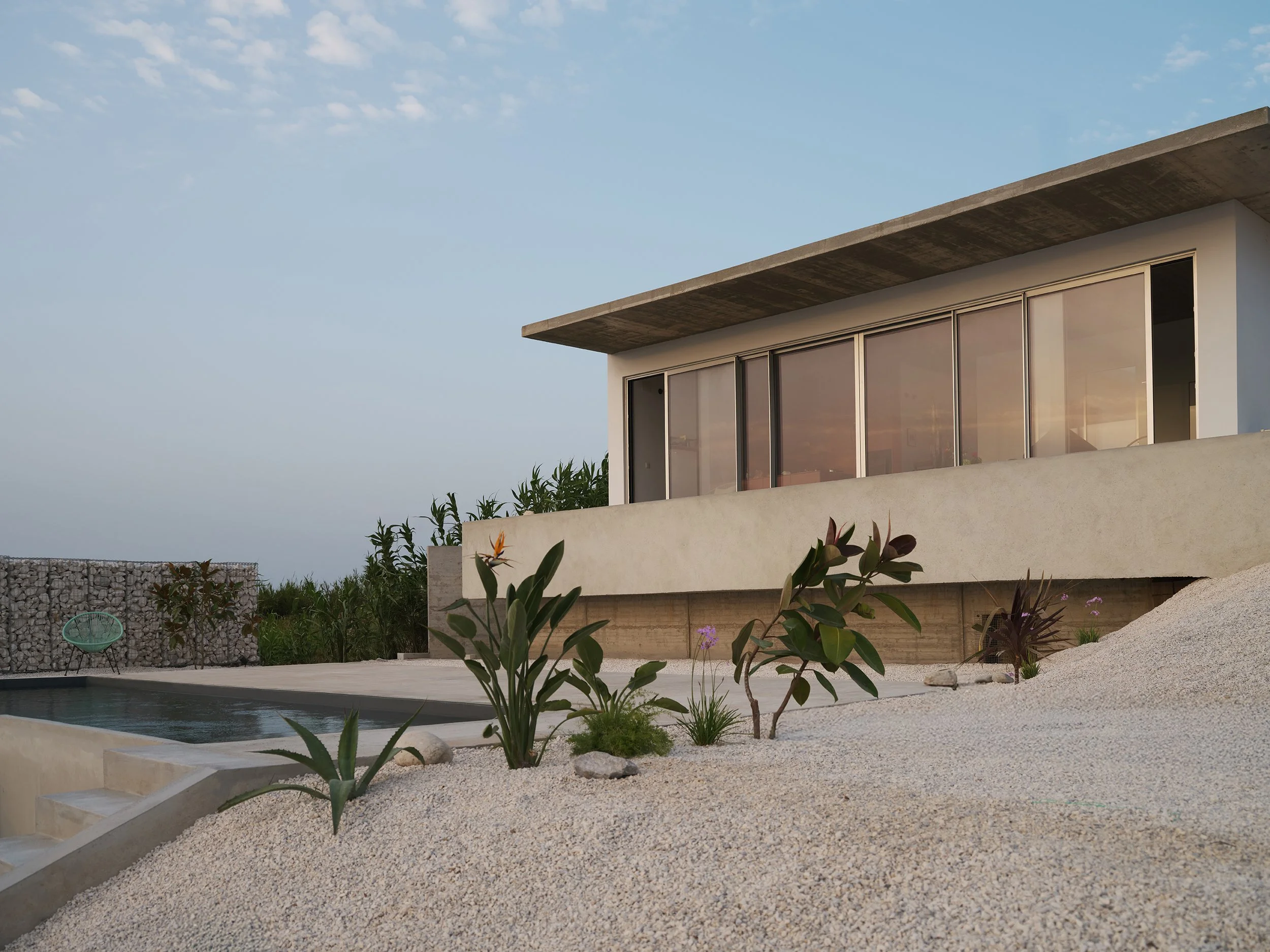 A swimming pool next to a concrete deck with two glasses of white wine, a pair of paddleball paddles with a yellow ball, and a woven mat, with green plants and a hillside in the background.
