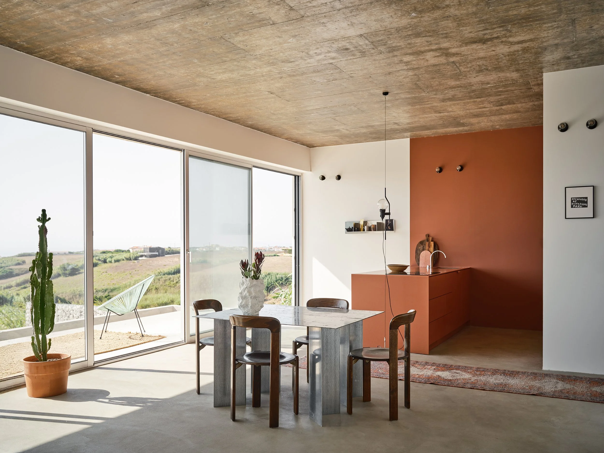 Modern kitchen with a terracotta-colored island, a wooden stool, and sunlight casting shadows on the beige floor, with a white wall and a backsplash with shelves in the background.