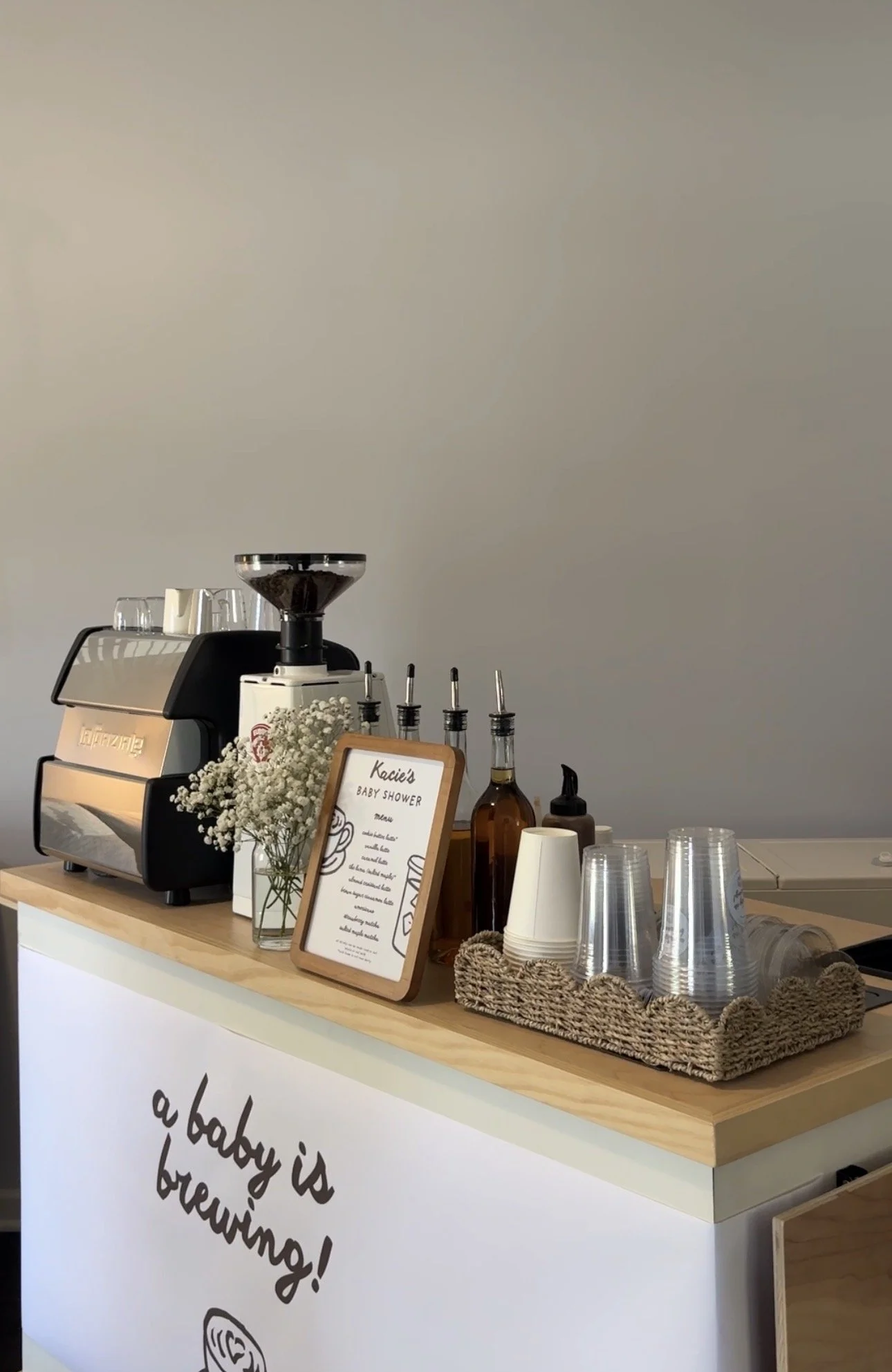 A mobile coffee bar setup with an espresso machine, various bottles, paper cups, and a sign for a baby shower.