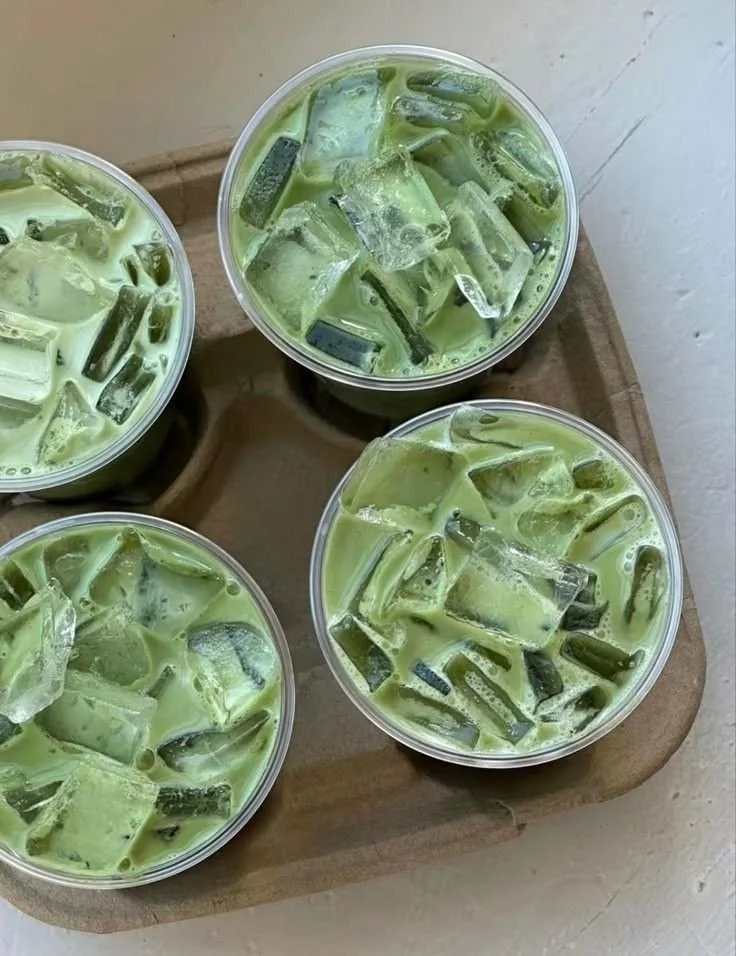 Four small clear plastic cups filled with a green matcha latte topped with ice cubes, placed on a wooden tray.