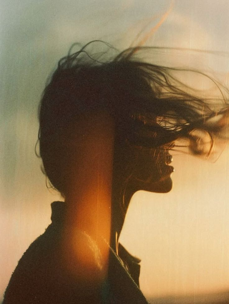 Silhouette of a woman with wind-blown hair, wearing large hoop earrings, against a sunset background