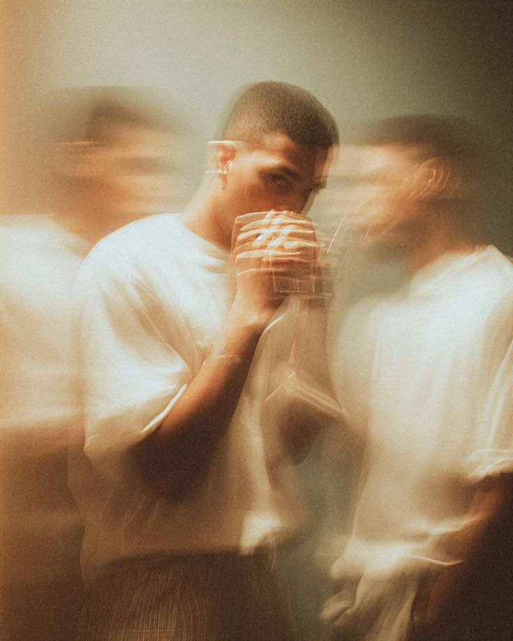 A young man with short hair holding a glass to his mouth, surrounded by blurred figures, creating a sense of motion or atmosphere.
