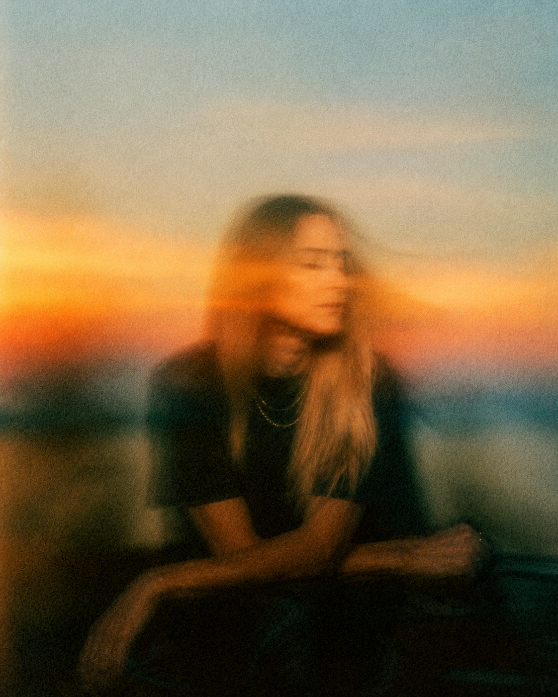 Blurry photo of a woman with long hair sitting outdoors at sunset with a colorful sky in the background.
