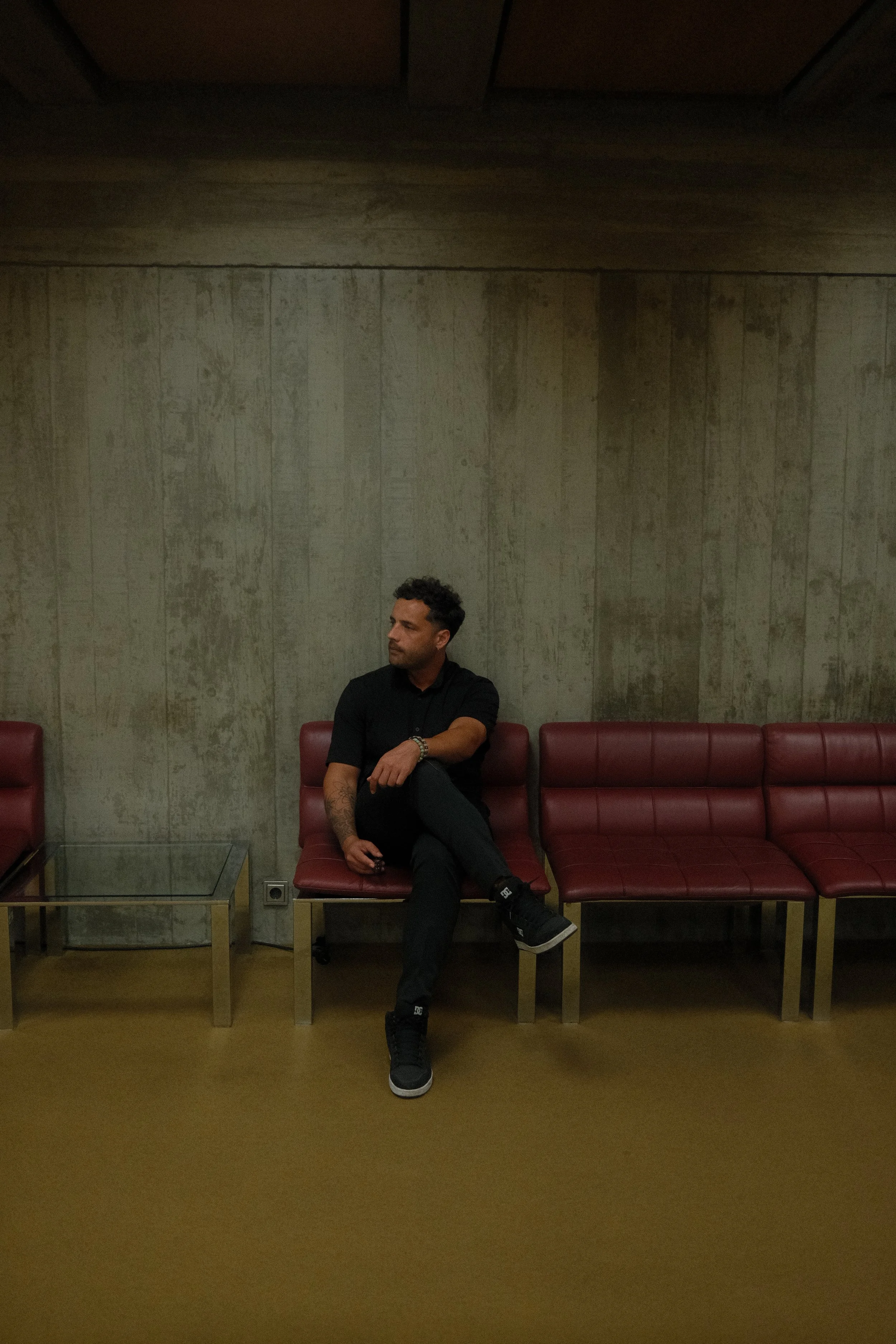 A man with dark curly hair and a light beard, dressed in black, sitting on a red cushioned bench in a room with a concrete wall and a wooden ceiling. He is looking to his right with his arms crossed, one ankle resting on his knee. There is a glass-topped table with a metal frame in front of him, and the floor is beige.