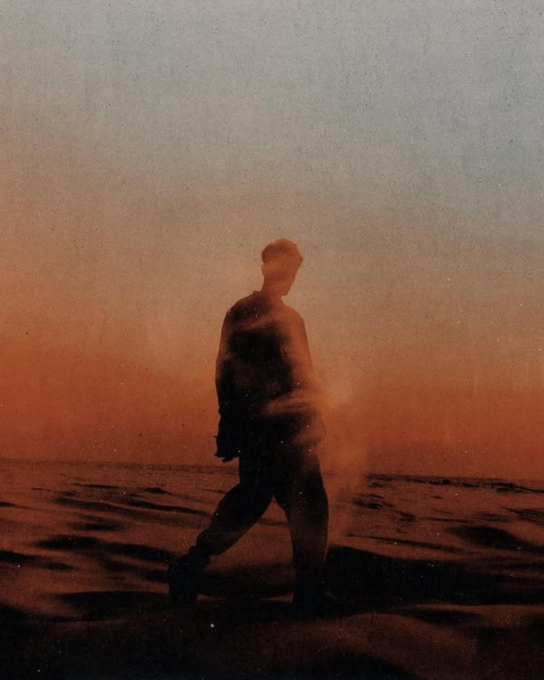 A person with short hair sitting on the beach at dusk with the ocean in the background and a cloudy sky, illuminated by an orange sunset.