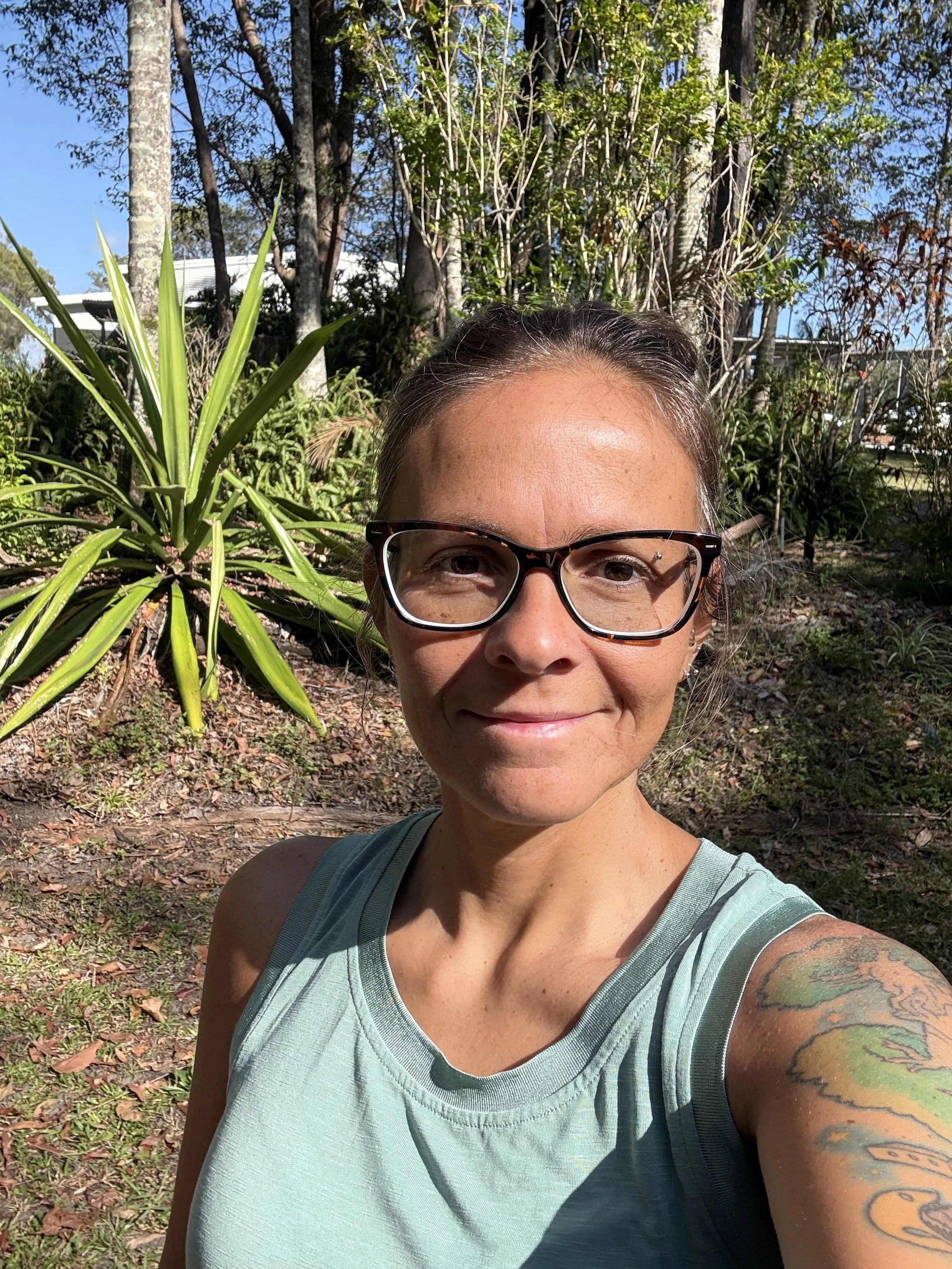 A woman wearing glasses and a light green sleeveless top taking a selfie outdoors, with trees and a sunny sky in the background.