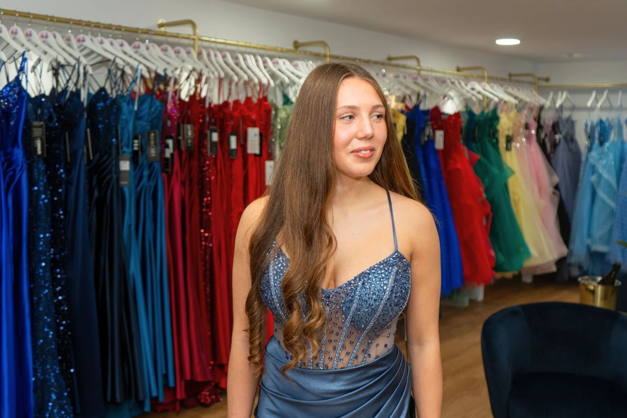 Young woman trying on a blue evening dress in a boutique with colourful gowns hanging on racks behind her.