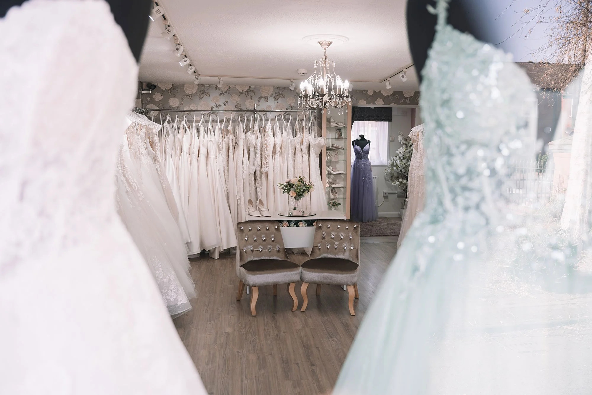 Inside a bridal boutique with wedding dresses hanging on racks, a mannequin in a blue dress, a chandelier, and a seating area with two upholstered chairs.
