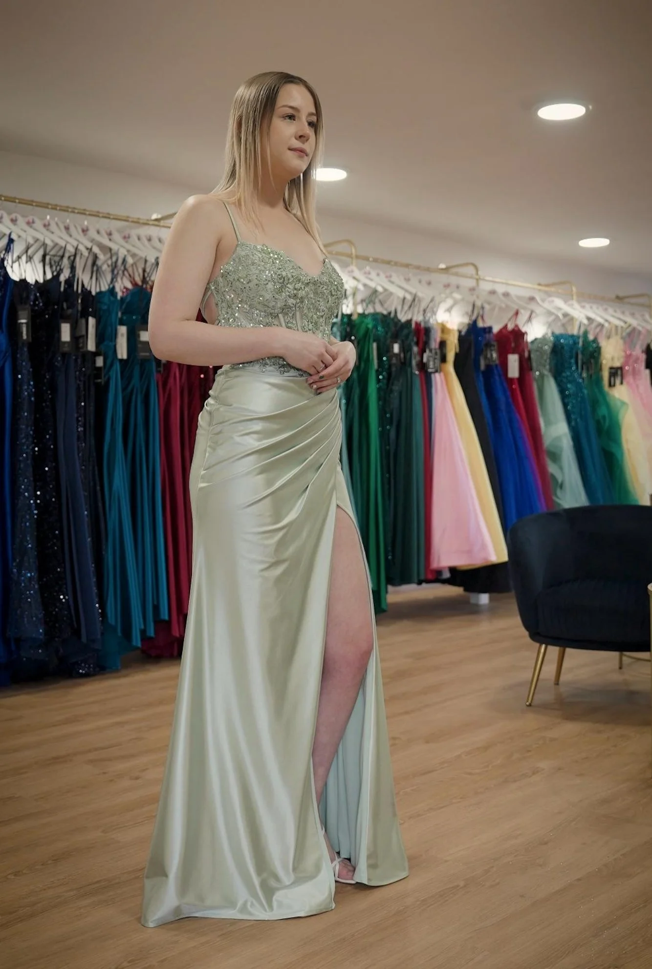 A woman trying on a long, elegant, light-coloured evening gown with a high slit at a dress boutique, surrounded by colourful dresses hanging on racks.