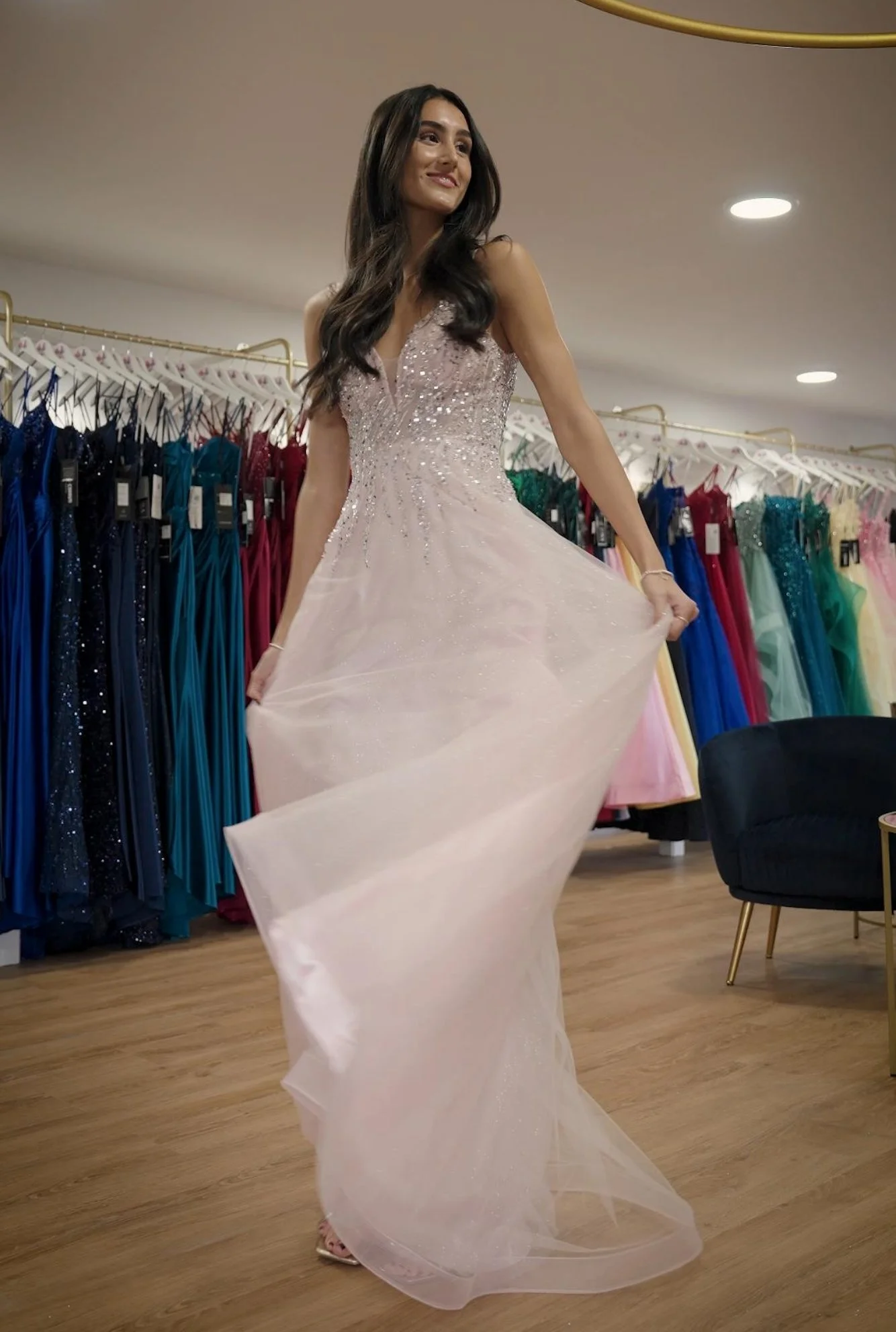 Woman trying on a light pink, glittery evening gown in a boutique with colourful dresses on hangers behind her.