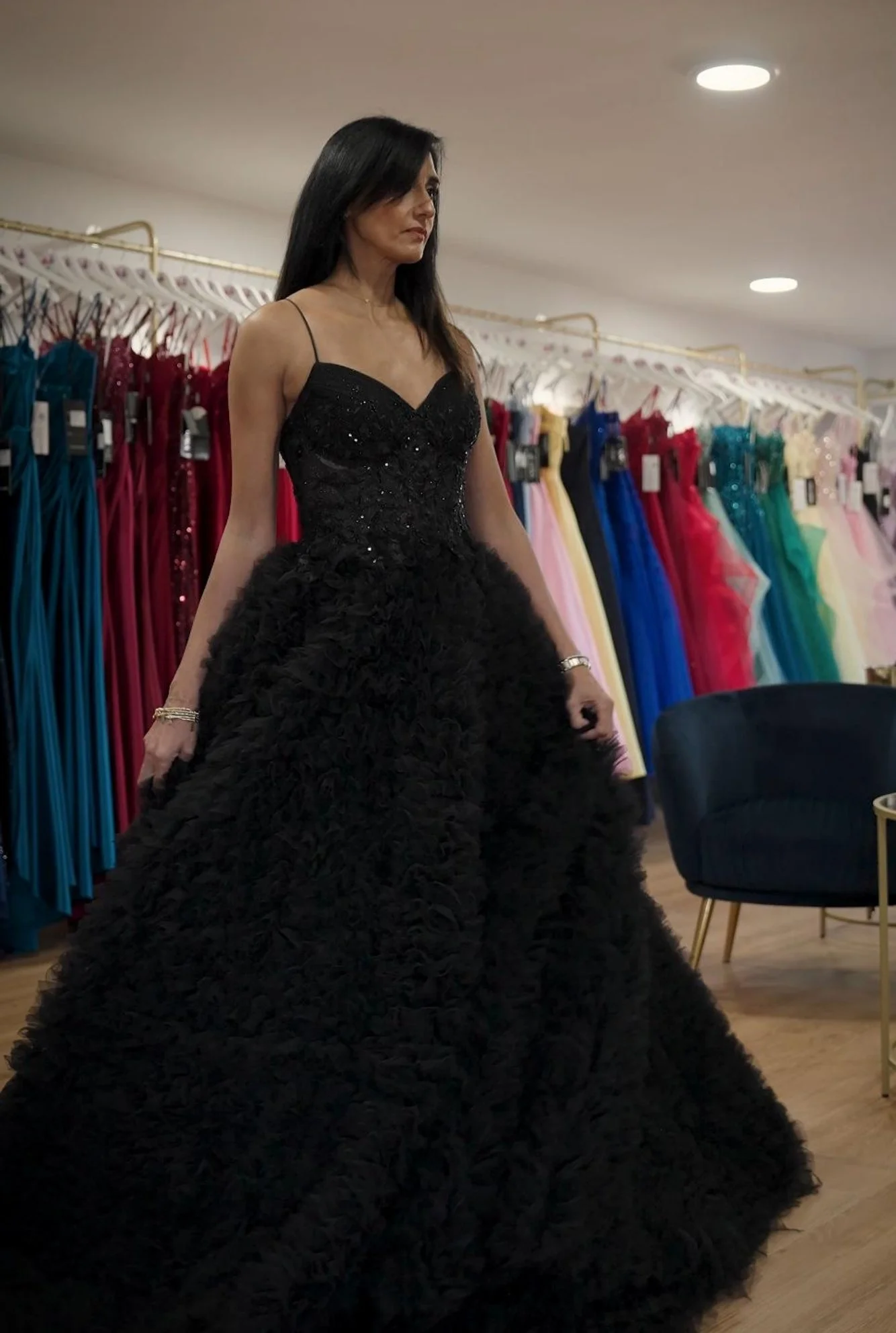 A woman in a black, strapless, beaded evening gown with a voluminous, textured skirt standing in a dress shop with colourful dresses on racks in the background.