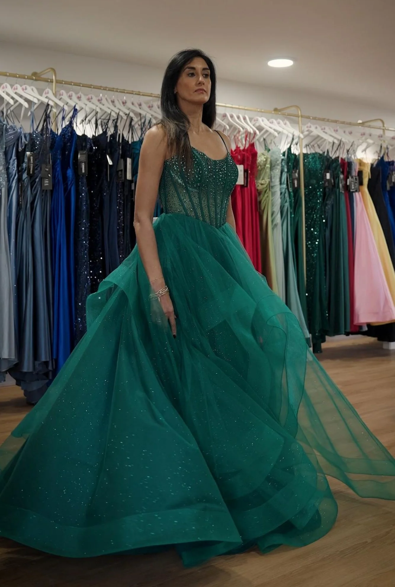 A woman trying on a green evening gown in a dress boutique, with colourful dresses hanging in the background.