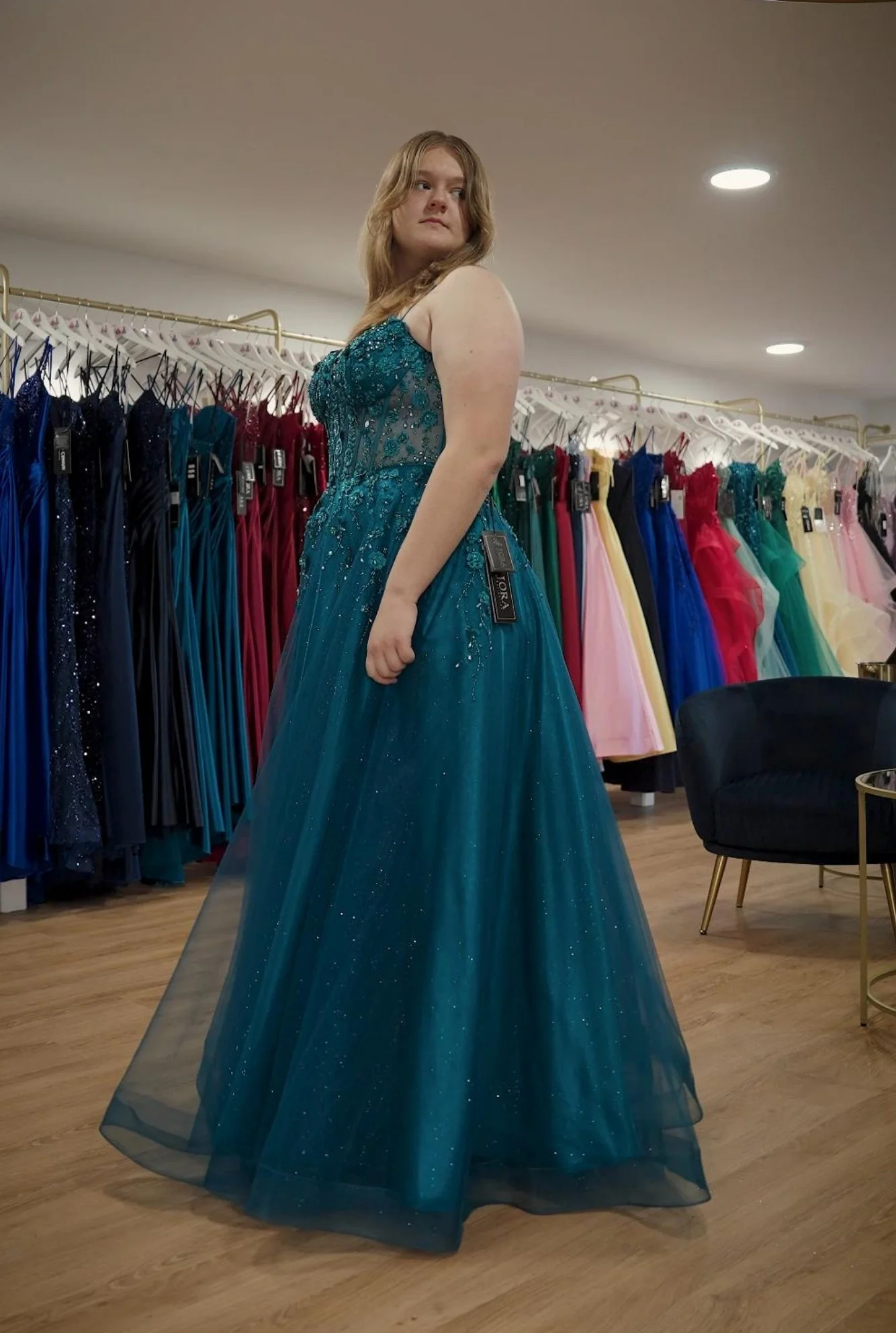 A woman trying on a teal evening gown with intricate beadwork and a full tulle skirt in a formal dress shop, surrounded by colourful dresses on racks.