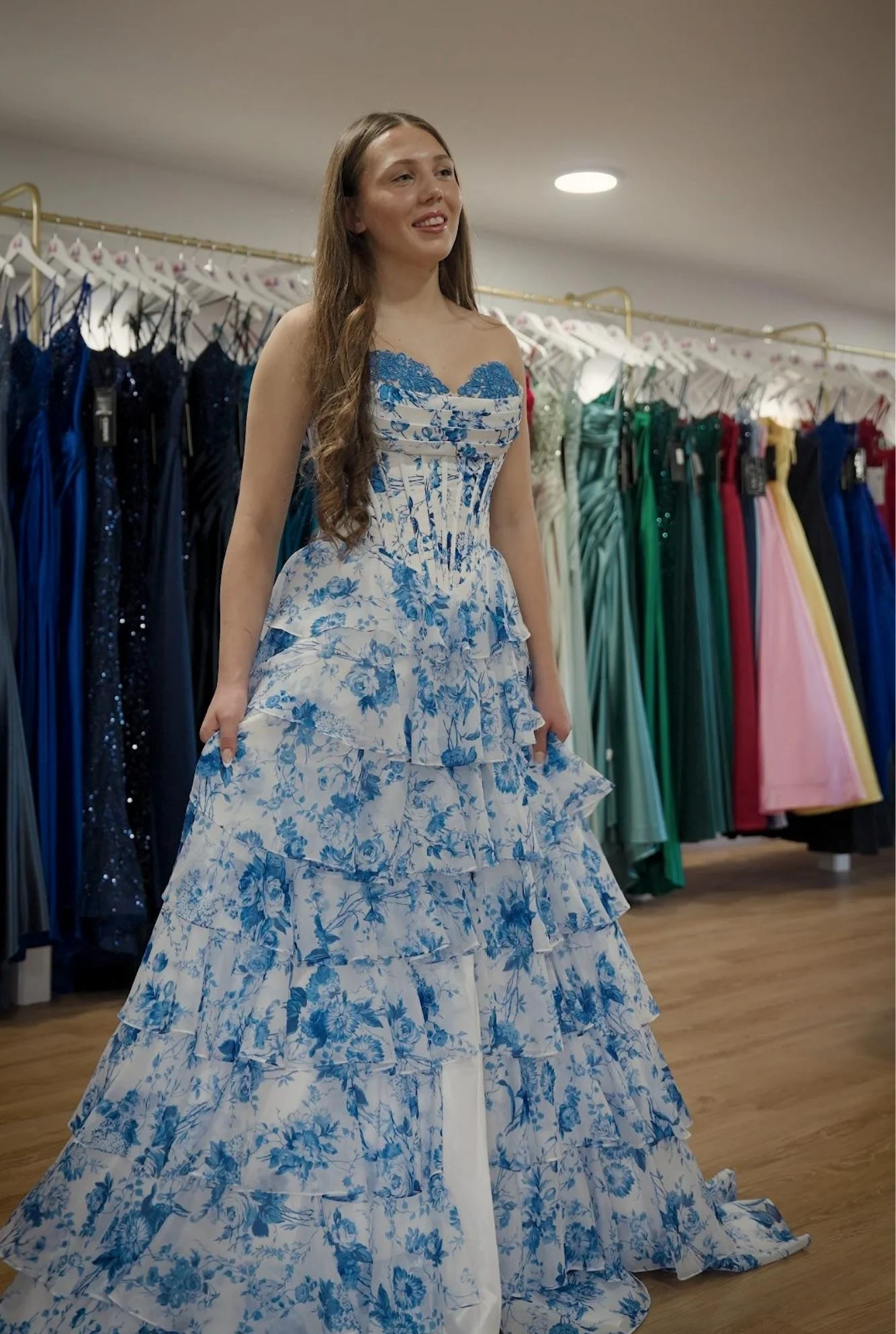 Woman trying on a white and blue floral prom dress in a formal wear store, with colourful dresses on racks in the background.
