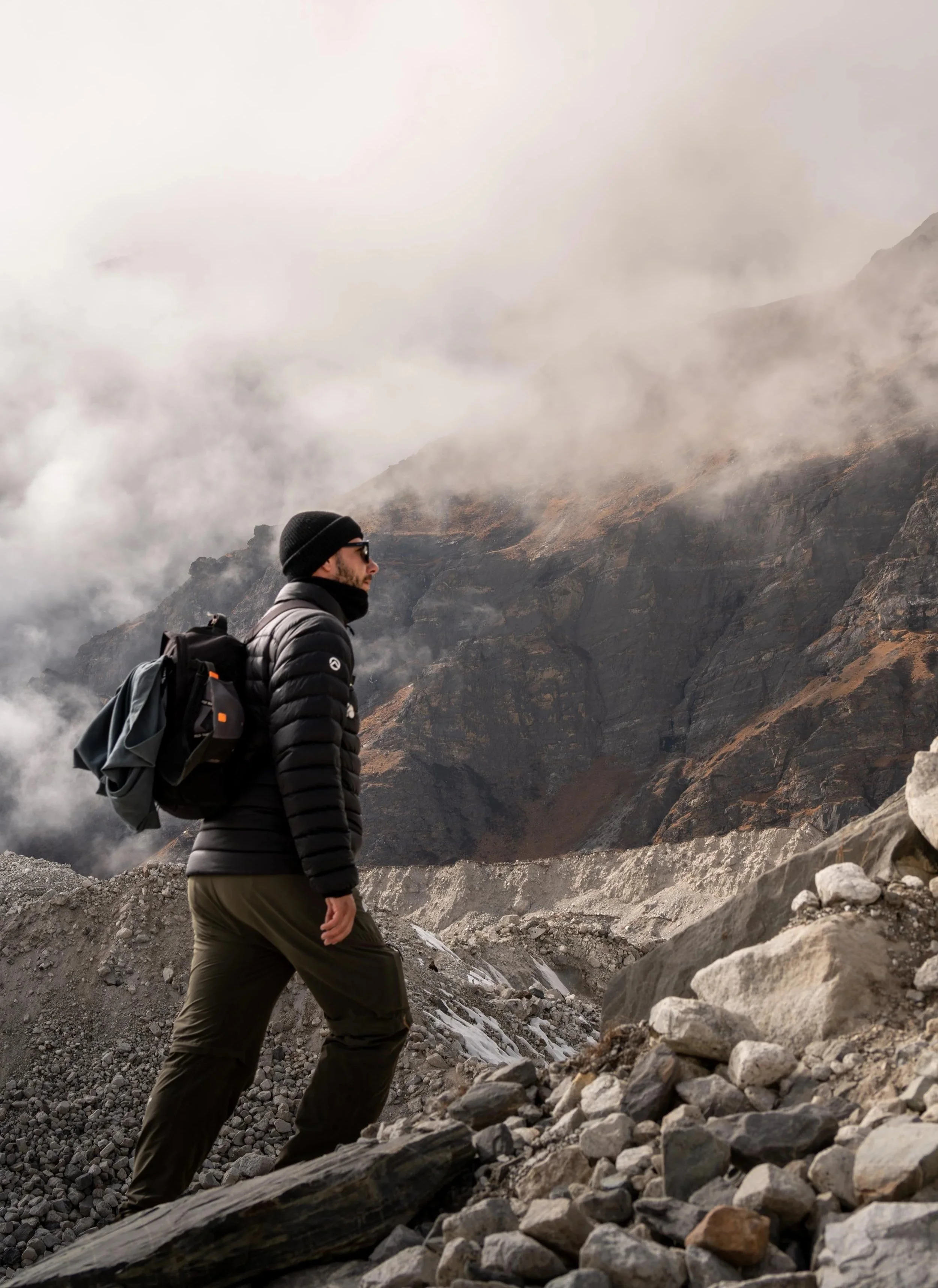 Un homme marche dans un paysage montagneux brumeux, portant un manteau noir, un bonnet noir, des lunettes de soleil et un sac à dos.