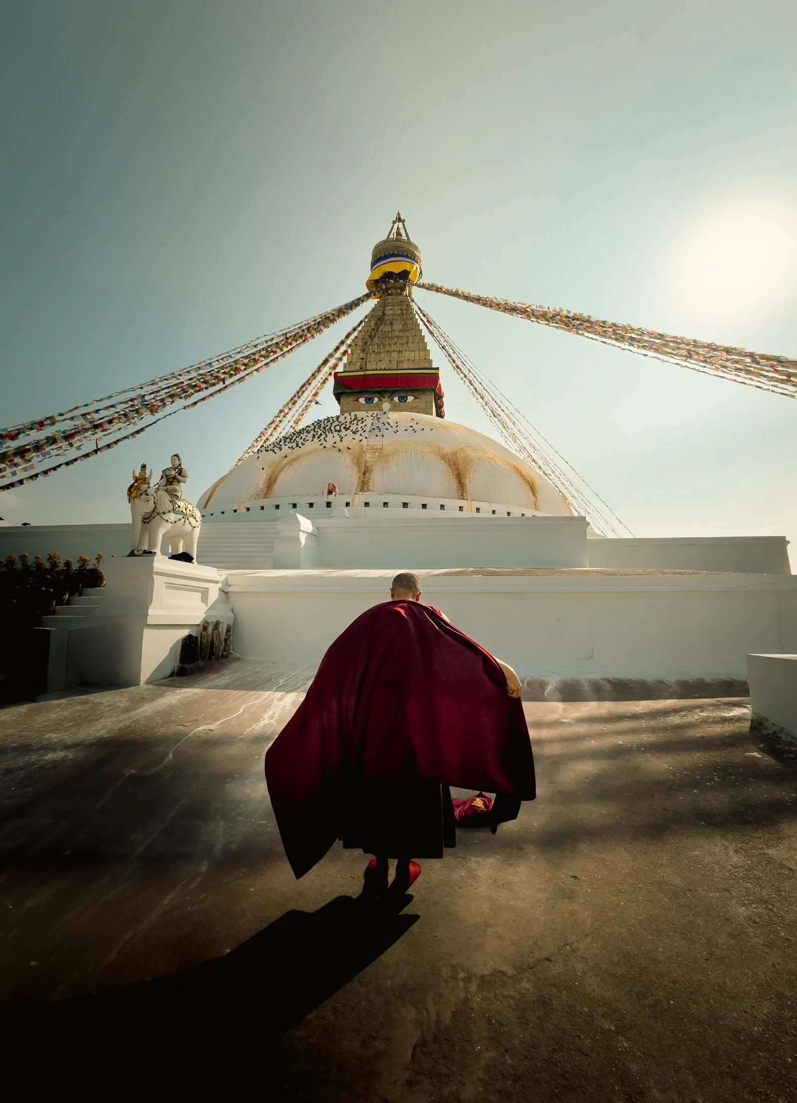 Un moine en robe rouge marchant devant la grande stupa de Boudha, décorée de drapeaux de prières colorés, dans un environnement en plein soleil.